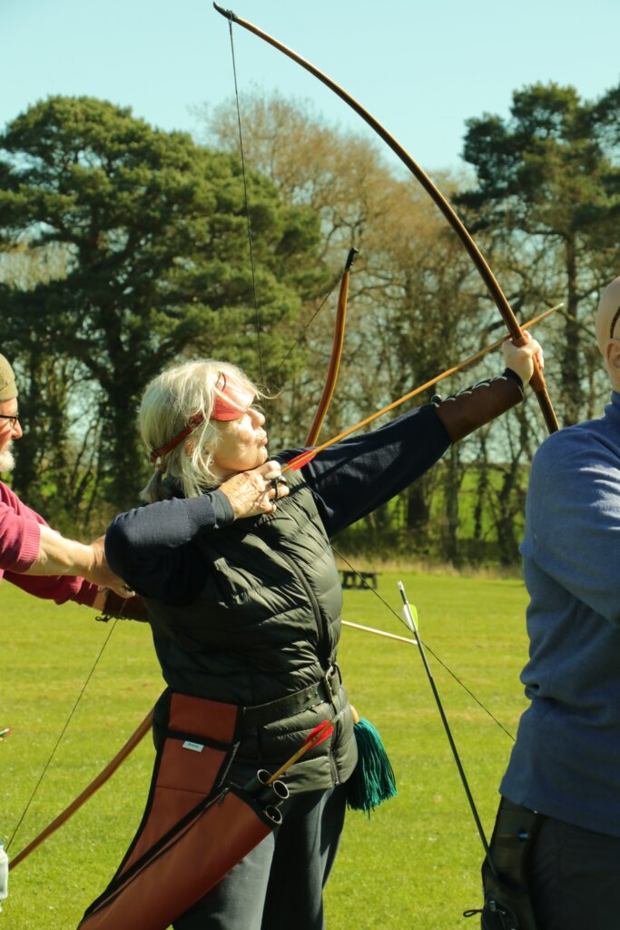 An archer at full draw with a longbow while a coach helps with her position
